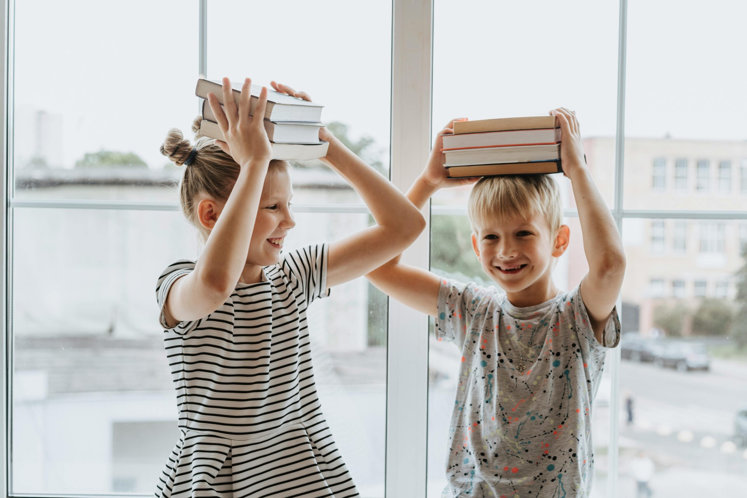 Two laughing siblings playfully balance books on their heads by a window, symbolizing fun learning.